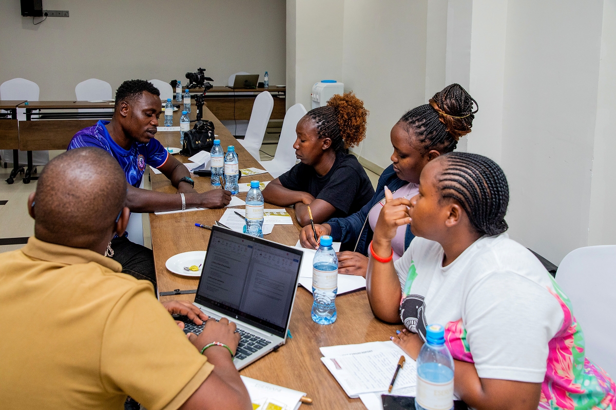 Five participants work together at a conference table with a laptop, notebooks, and printed documents.