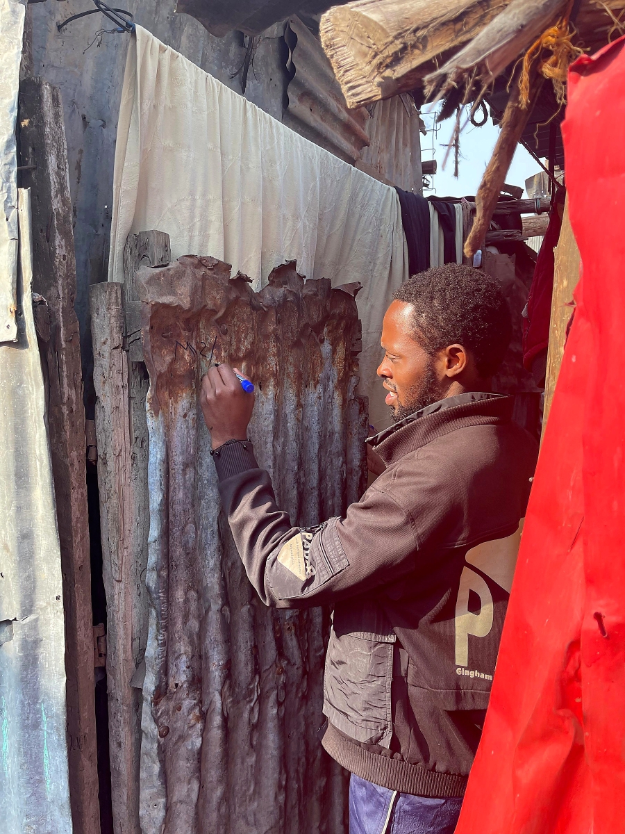 A man writes a number on a corrugated metal wall in a narrow settlement passage.