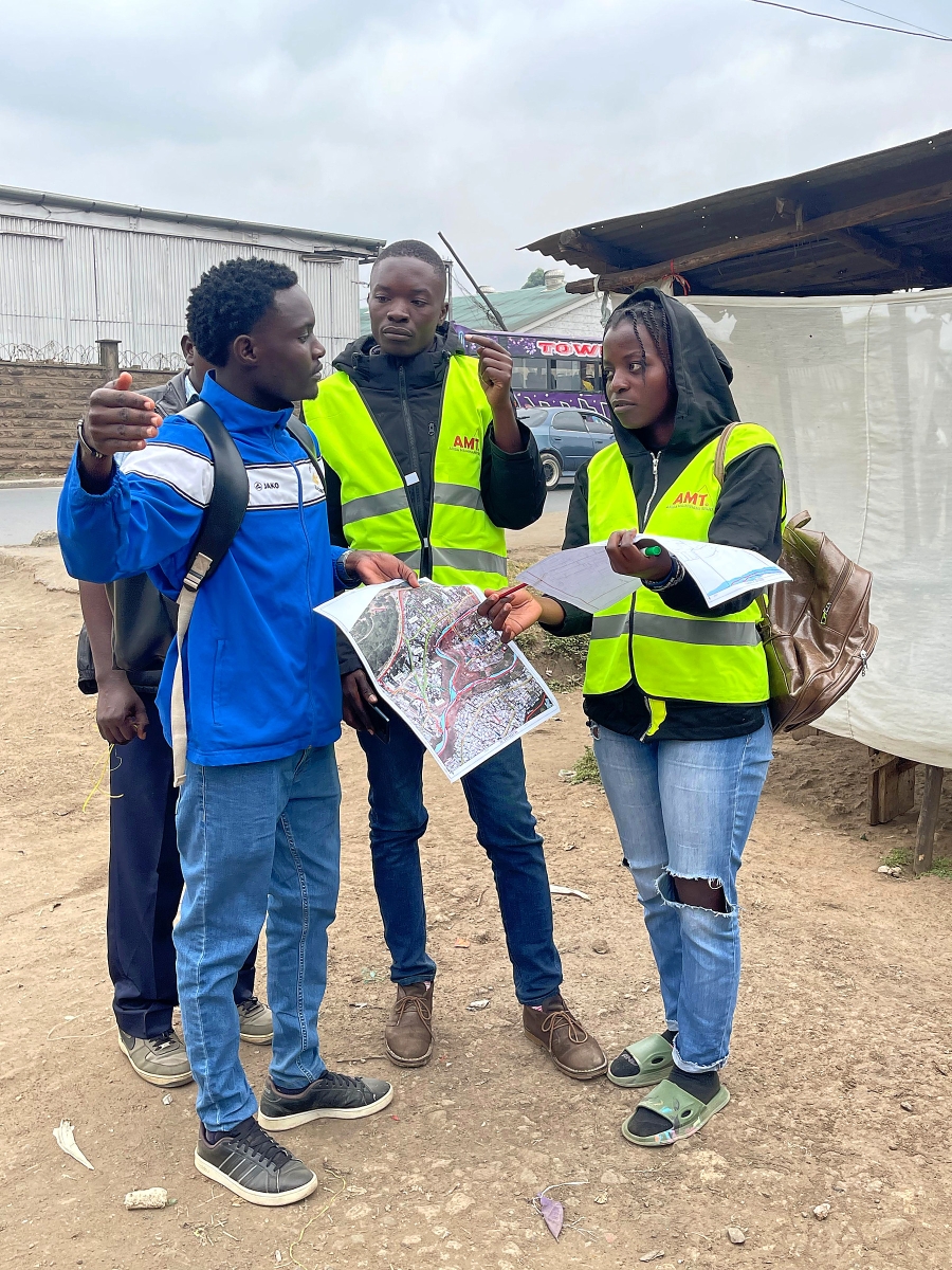 Three researchers in reflective vests compare printed maps beside a road as one person gestures toward the area.